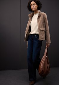 Brązowa kurtka ze sztucznego futra, kremowy sweter, ciemnoniebieskie dżinsy oraz brązowa skórzana torba na ramię. Miękka tekstura, casualowy krój i minimalistyczne detale.