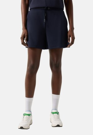 Persoon draagt marineblauwe shorts met trekkoord, witte sokken en witte sneakers met groene veters, staand tegen een effen achtergrond.