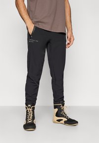 Calças desportivas pretas com pernas afuniladas e bolsos laterais com fecho, combinadas com sapatos altos pretos e bege com painéis de malha e atacadores.