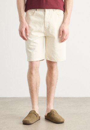 Beige denimshorts med avslappet passform, midt på låret, og diskrete lommer. Kombinert med brune semskede klippere på en nøytral gulv.