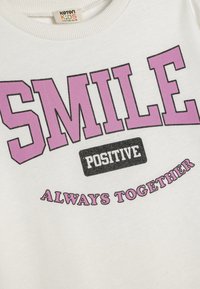Witte sweatshirt met een ronde halslijn, voorzien van vetgedrukte paarse tekst: "SMILE," "POSITIVE," en "ALWAYS TOGETHER" in zwarte accenten.