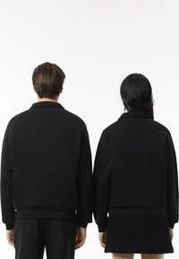 Sudaderas negras de corte relajado, con un cuello de camisa y puños y dobladillo de canalé, llevadas por dos personas vistas desde atrás.