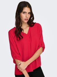 Rode blouse met korte mouwen en een V-hals, met opgerolde mouwen en een ontspannen pasvorm. Draagt over zwarte broek, effen achtergrond.