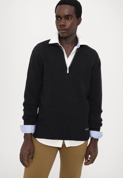 Svart zip-necksweater i stickad kvalitet, med ribbade ärmslut och nederkant, lagd över en vit skjorta med blå accent ärmslut.
