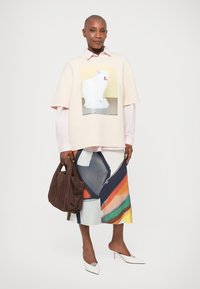 Svetlo roza oversized sweatshirt s grafiko mačke, oblečeno čez belo srajco; pisana vzorčasta krila; rjava torba; bele pete.