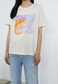 Persona vistiendo una camiseta color crema con el texto "Paz y Amor" en púrpura, naranja y azul sobre un fondo de patrón paisley, combinada con unos jeans claros rasgados.