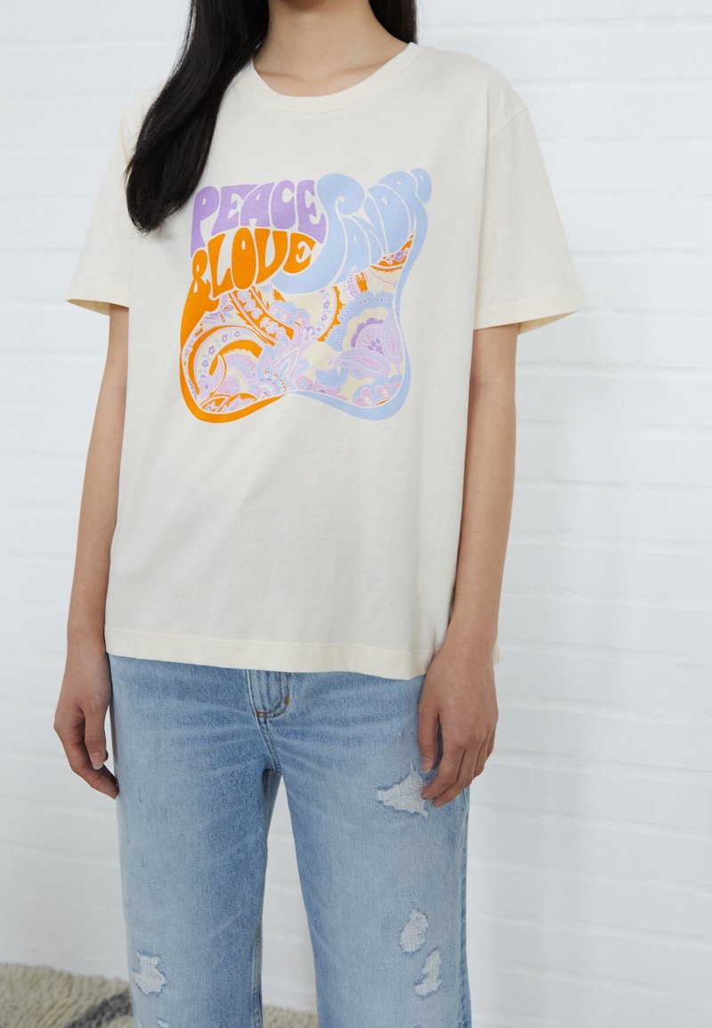 Persona vistiendo una camiseta color crema con el texto "Paz y Amor" en púrpura, naranja y azul sobre un fondo de patrón paisley, combinada con unos jeans claros rasgados.