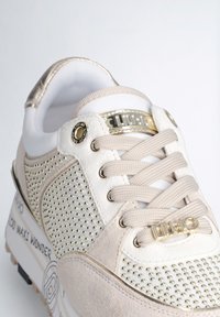 Sneaker beige e bianco con accenti metallici, texture punteggiata e lacci. Presenta un logo sul lato e una suola ammortizzata.
