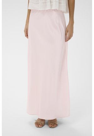 Kobieta ubrana w długą, jasnoróżową spódnicę, do której dobrała beżowe sandały z odkrytymi palcami oraz biały top ozdobiony koronką, stoi na tle jednolitego tła.