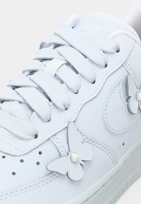 Nærbilde av hvit lær sneakers med tykke lisser og dekorative sommerfuglfasongede lærlapper festet på siden.