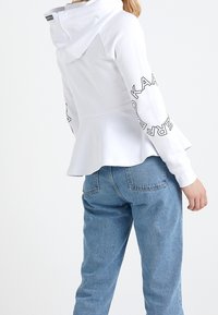 Felpa bianca con peplum, dotata di cappuccio, stampa del logo sulle maniche, abbinata a jeans azzurri chiari. Il tessuto sembra morbido, con una trama liscia.