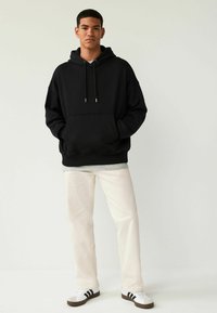 Sort oversized hoodie med kængurulomme, justerbar snor, parret med lyse lige bukser og sort-hvide sneakers.