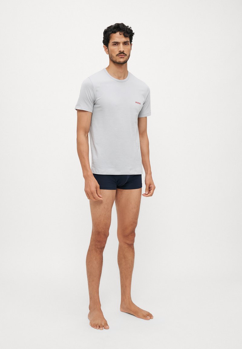 Lys grå bomull t-skjorte med rund hals og liten rød "HUGO" logo, kombinert med marineblå shorts. Enkel, tettsittende design.
