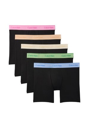 Uomo senza maglietta in piedi scalzo, indossa boxer neri con una cintura rosa Calvin Klein, aggiustando la fascia in vita con entrambe le mani.