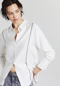 Camicia bianca con bottoni e righe verticali nere, caratterizzata da una texture sottile, colletto e maniche arrotolate. Abbinata a denim grigio.