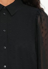 Blouse noire en tissu texturé avec des motifs à pois en relief, manches longues en organza, et un col classique avec fermeture à boutons.