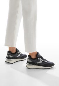 Black and gray sneakers with a textured upper, metallic accents, and a cushioned sole, paired with loose white cropped pants.