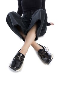 Mocasines de charol negro con un acento de hebilla en tono dorado, que presentan una suela de goma beige en contraste y una textura suave y brillante.