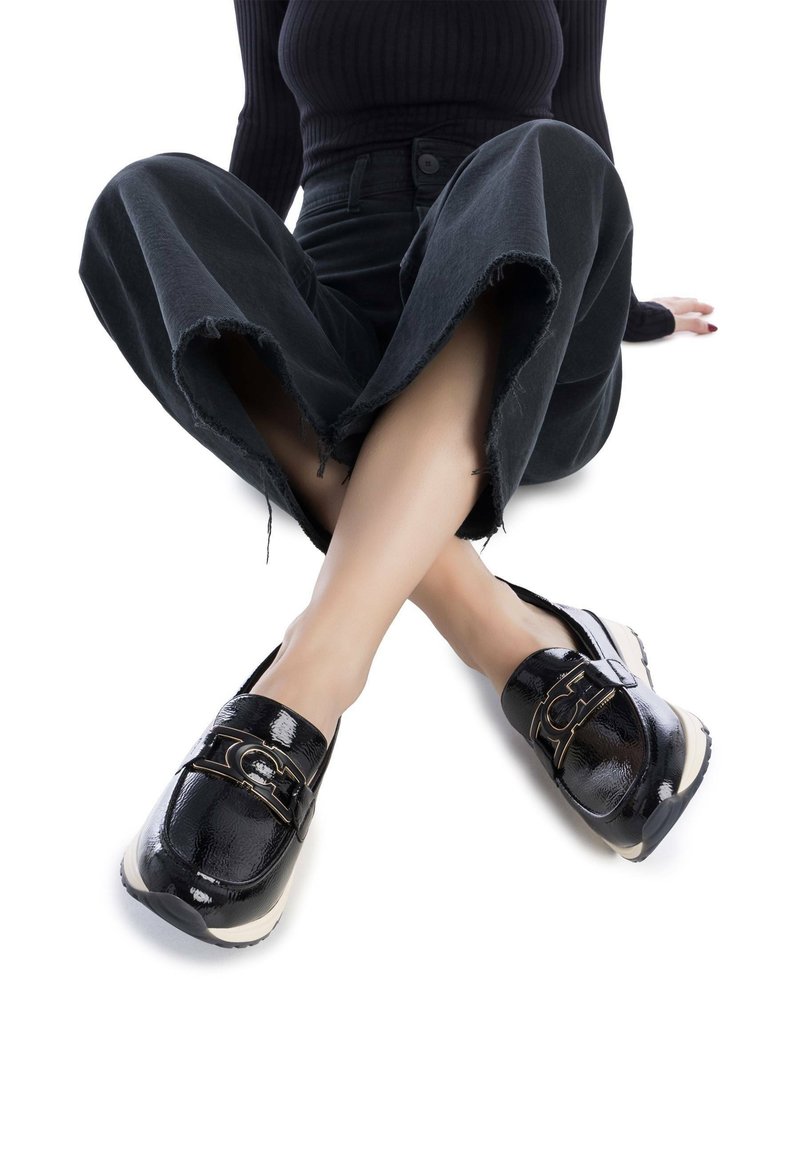 Mocasines de charol negro con un acento de hebilla en tono dorado, que presentan una suela de goma beige en contraste y una textura suave y brillante.