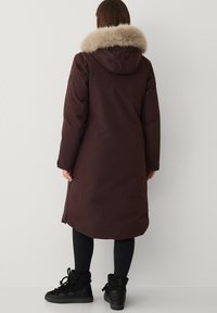 Lange bordeauxrode parka met een capuchon van nepbont. Heeft een getailleerde pasvorm, met ritsen sluitende zakken en wordt gedragen met zwarte enkellaarzen.