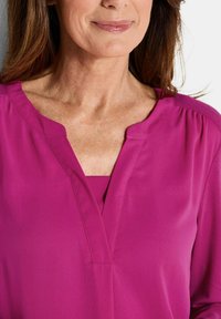 Magenta Bluse mit V-Ausschnitt und gerafften Schultern, aus glattem Stoff. Mit verdeckt verarbeiteten Nähten entlang des Ausschnitts.