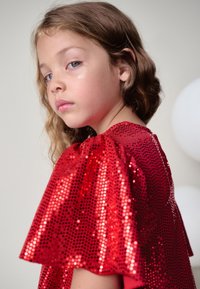 Rød paljettop med pufærmer, der har en blank tekstur og rund halsudskæring. Modellen har bølget hår og neutral makeup.