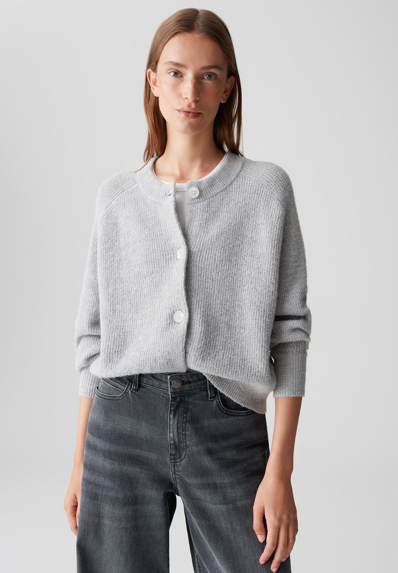 Femme portant un cardigan boutonné gris clair sur un haut blanc avec un jean gris foncé, se tenant devant un fond uni.
