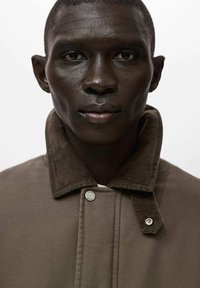 Giacca marrone con collo in velluto a coste, con bottoni a pressione in metallo e tessuto strutturato, primo piano dell'area del collo su sfondo bianco.