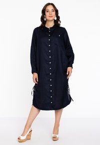 Marineblaues Leinenhemdkleid mit Knopfleiste, seitlichen Bindebändern und einer einzelnen Brusttasche. Es hat lange Ärmel und einen lässigen Schnitt.