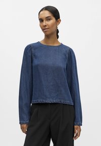 Tamni plavi traper gornji dio s dugim rukavima, okruglim izrezom i nabranim ramenskim šavovima. Rub i manžete imaju sirove rubove za teksturirani završetak.