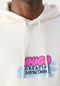 Sweatshirt crème avec une capuche à cordon. Présente un logo coloré avec des accents roses et bleus indiquant "HUGO CREATIVE DEPARTMENT". Texture douce.