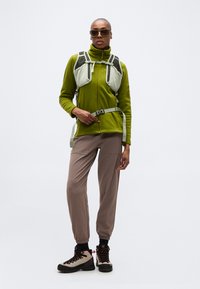 Άτομο που φοράει πράσινο φλις μπουφάν με φερμουάρ, μπεζ παντελόνι πεζοπορίας, μποτάκια πεζοπορίας, γυαλιά ηλίου και γκρι τιράντες σακιδίου πλάτης, στέκεται μπροστά σε λευκό φόντο.