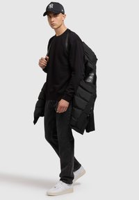 Joven vestido con sudadera negra, jeans negros, zapatillas blancas, una gorra negra, y llevando una chaqueta acolchada negra sobre un hombro.