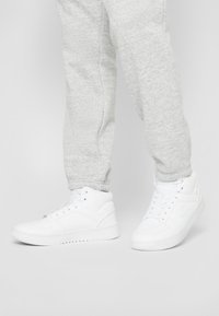 Sneaker alte bianche realizzate in materiale sintetico liscio, con colli imbottiti, chiusura con lacci e suole in gomma testurizzata.