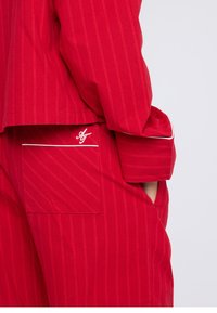 Punaraidallinen asu, jossa tasku valkoisella reunuksella ja kirjailtu logo. Kangas näyttää sileältä ja istuu räätälöidysti.