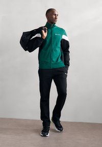 Tuta verde e nera con dettagli bianchi, caratterizzata da una giacca con zip e pantaloni con elastico in vita. Sneakers nere con dettagli gialli.