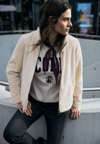 Kremowa kurtka z futerka ekologicznego z zamkiem z przodu, w połączeniu z jasną koszulą z ciemnym napisem i grafiką oraz ciemnoczerwoną szalikiem.