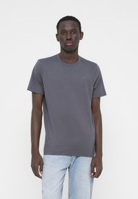 Pilkas trumpomis rankovėmis marškinėlis, pagamintas iš medvilnės mišinio audinio, su apvalia iškirpte ir mažos logotipo detales ant krūtinės, derinamas su šviesiai mėlynais džinsais.