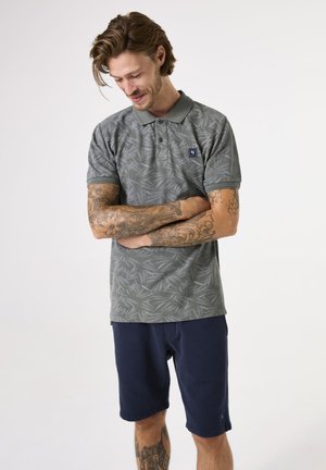 Polo gris avec un motif de feuilles, à manches courtes et avec un col. Porté avec un short bleu marine. Comprend un petit écusson logo sur la poitrine.