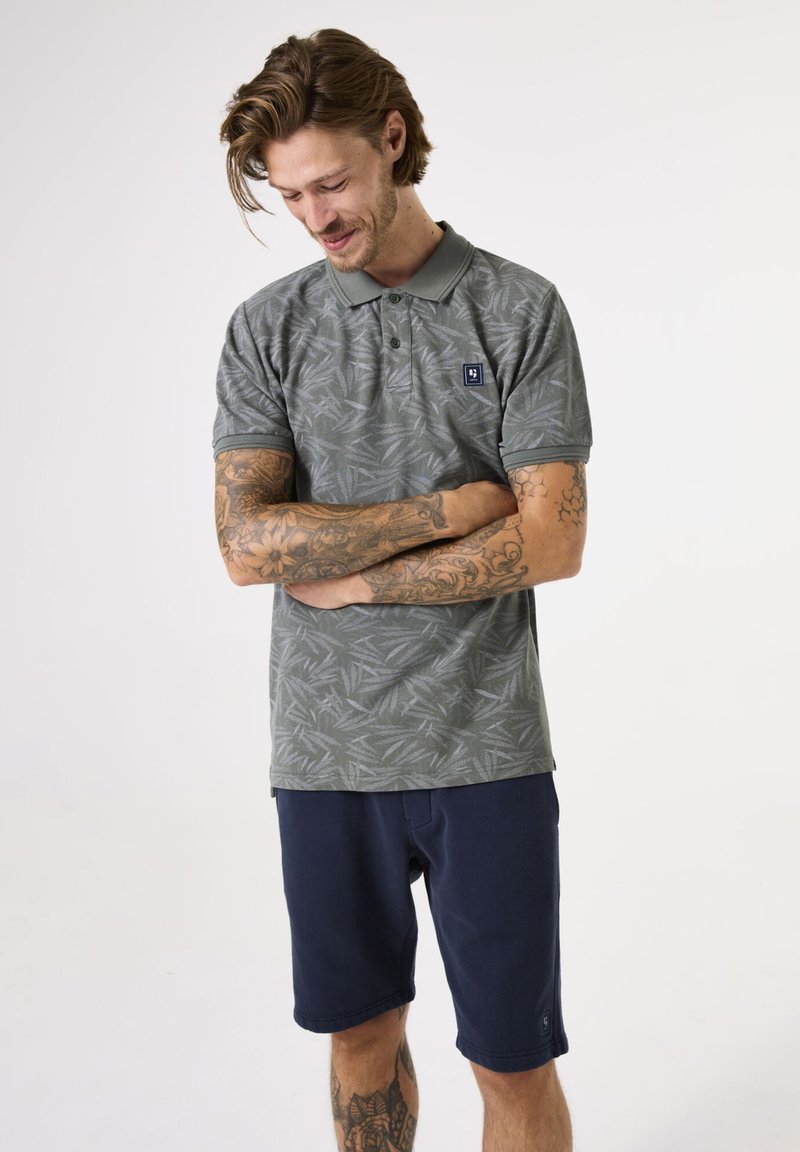 Grijze poloshirt met bladpatroon, korte mouwen en een kraag. Draagt bij navy shorts. Heeft een klein logo op de borst.