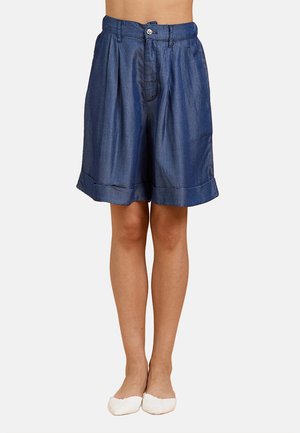Pantaloncini blu a vita alta plissettati con orli risvoltati indossati da una persona in piedi con gambe nude e ballerine bianche.