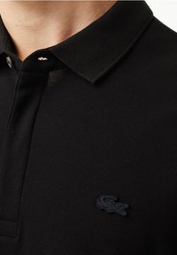 Schwarzes Polohemd mit strukturiertem Stoff, versehen mit einem klassischen Kragen und einem marineblauen, gestickten Logo auf der Brust. Einfarbig, ohne Muster.