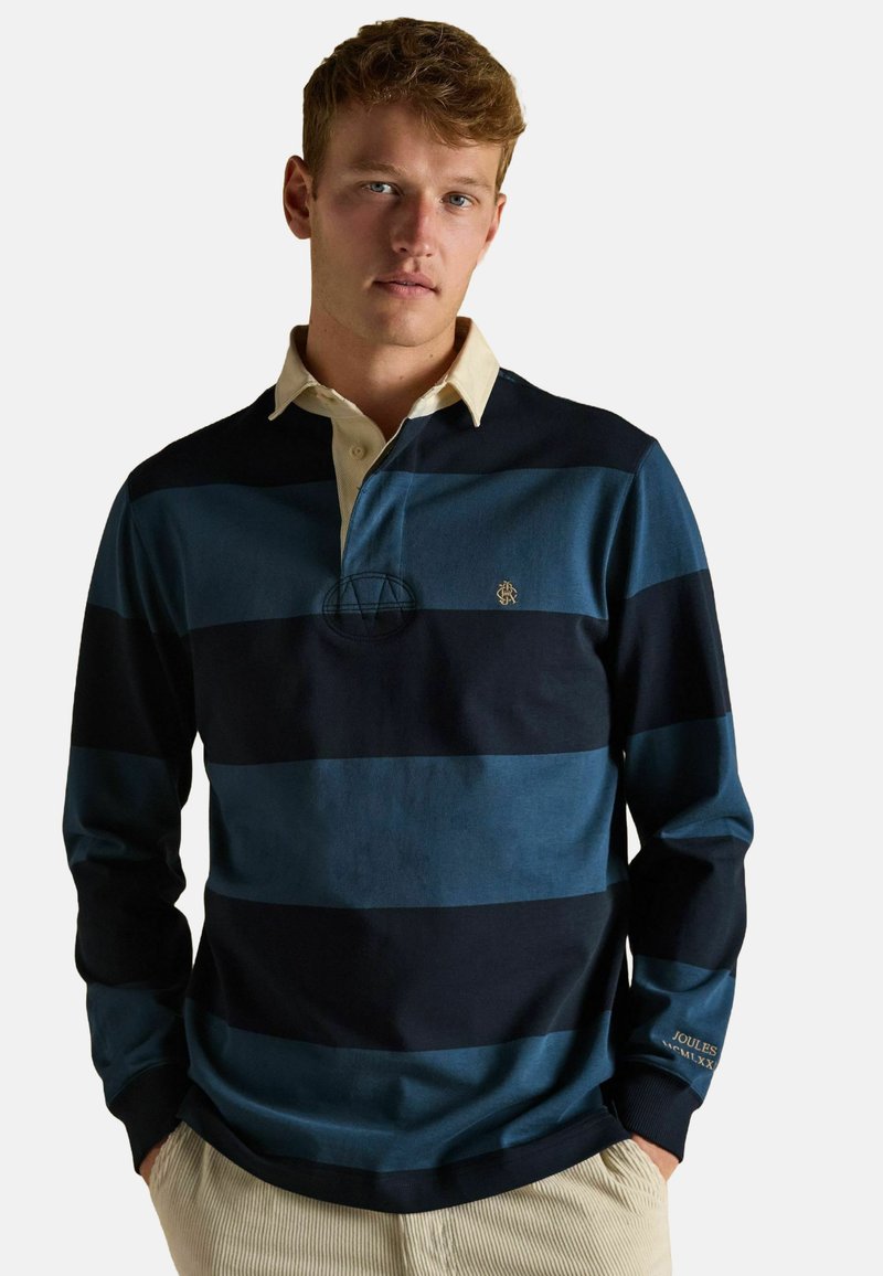 Junger Mann mit kurzen blonden Haaren, der ein blau-schwarz gestreiftes Rugby-Shirt mit beigem Kragen und hellbeigen Hosen trägt, mit den Händen in den Taschen.