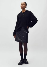 Sort strikket sweater med krave og tre knapper, parret med en sort lædernederdel med hvide nitter. Modellen har sorte strømpebukser og sko på.