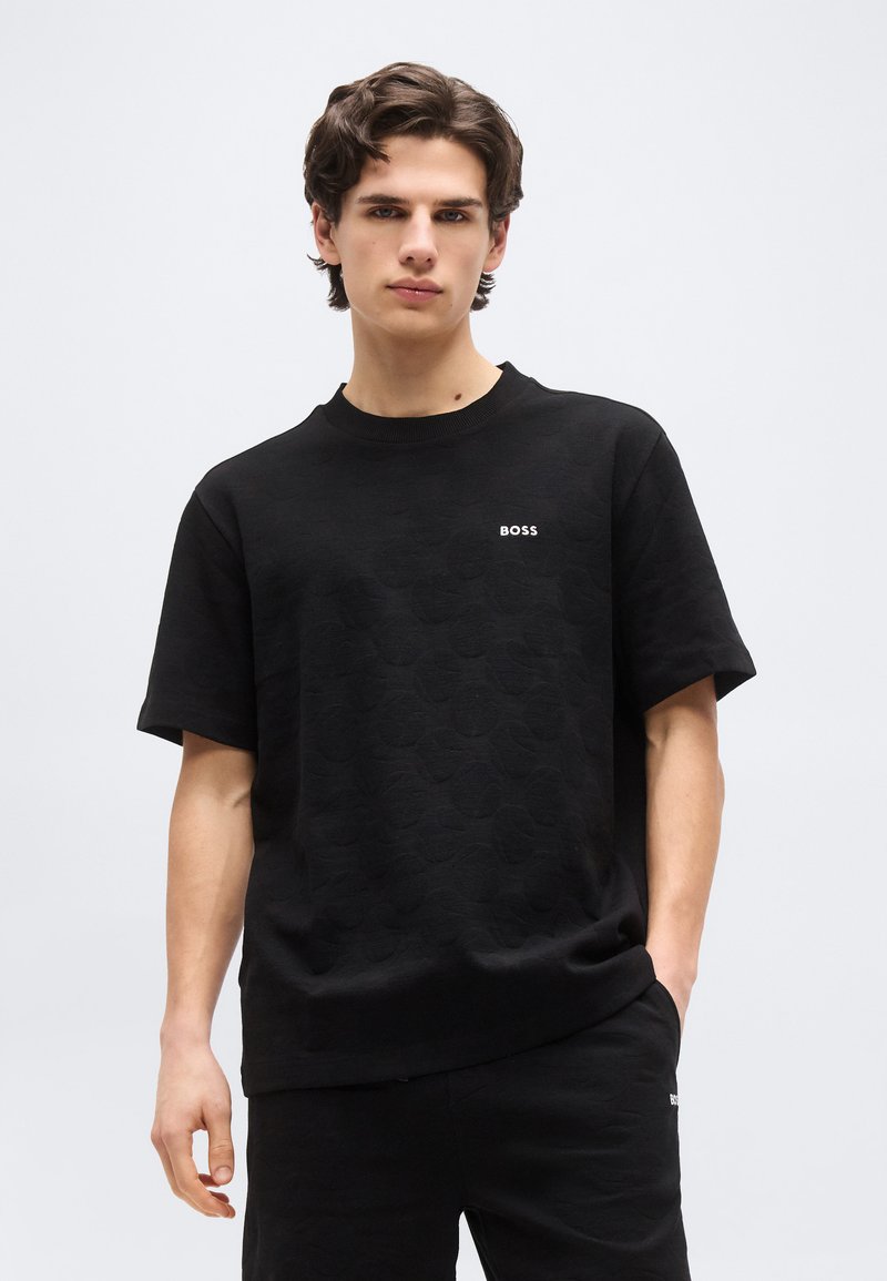 Νεαρός άνδρας που φοράει μαύρο κοντομάνικο πουκάμισο και αντίστοιχο μαύρο παντελόνι, στέκεται με το ένα χέρι στην τσέπη, μπροστά σε απλό φωτεινό υπόβαθρο.