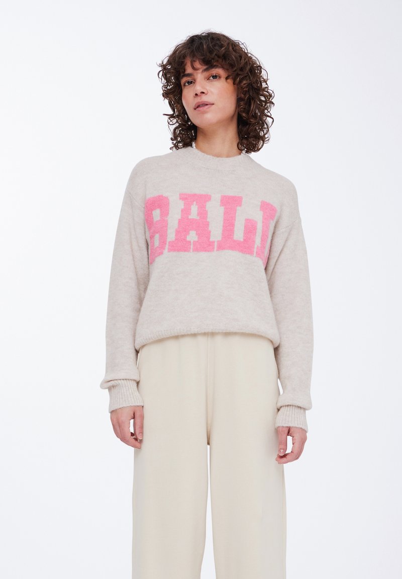 Beige fuzzy sweater med "BALI" i store pink bogstaver. Har en rund halsudskæring og ribbede manchetter. Sammen med højtaljede cremede bukser.