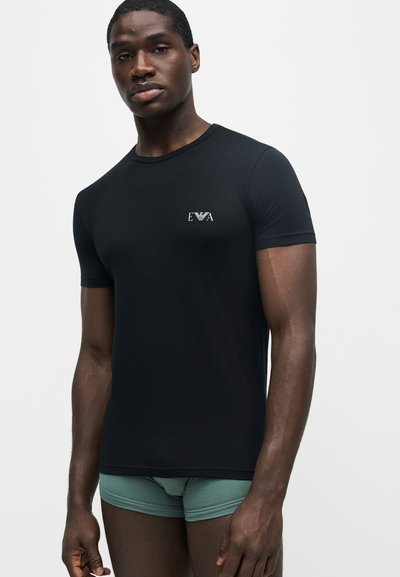 Άνδρας που φοράει εφαρμοστό μαύρο T-shirt με μικρό λευκό λογότυπο στο στήθος και ανοιχτοπράσινο εσώρουχο σε απλό φόντο.