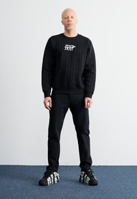 Sort strikket sweatshirt med lodrette striber og "JUST DO IT" logo, parret med sorte bukser og sorte sneakers med hvide accenter.