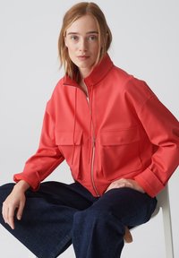 Rote, übergroße Jacke mit hohem Kragen, zwei Fronttaschen und einem Reißverschluss. Glatte Textur mit entspannter Passform, kombiniert mit dunkelblauen Hosen.