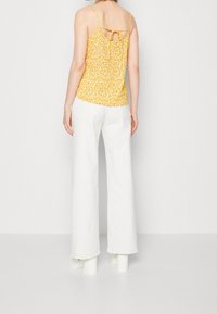 Camiseta floral amarilla con tirantes finos y detalle de lazo en la parte posterior, combinada con pantalones anchos blancos y zapatos de tacón chunky.